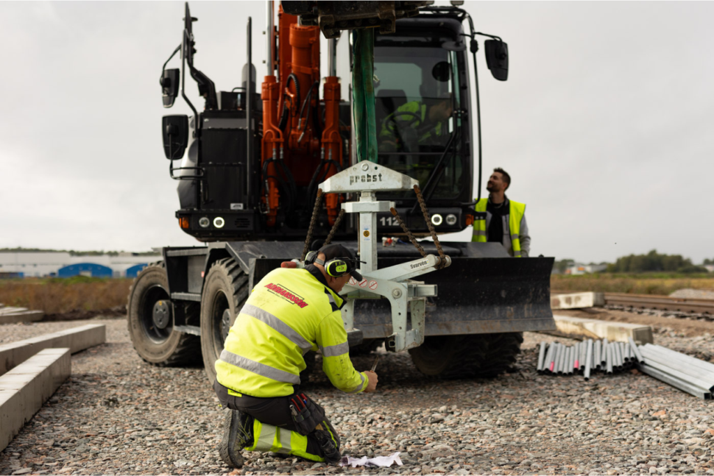 Arbetare i varselkläder monterar utrustning framför en grävmaskin på en byggarbetsplats, kopplat till avtal om jordhantering i Örebro, medan en kollega står i bakgrunden.